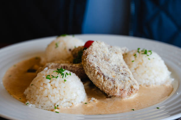 A plate featuring breaded meat served with rice balls and a creamy sauce, garnished with fresh herbs and a small tomato slice. The dish is presented on a white plate, offering a hearty and flavorful meal.