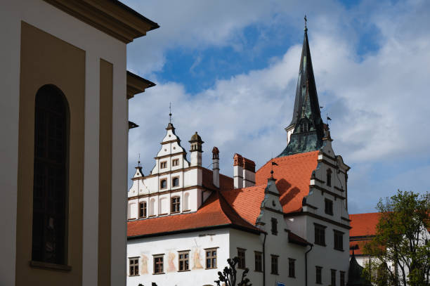 A historic church with a tall spire and an adjoining building stand beside a cobblestone square under a partly cloudy sky. The architecture features traditional elements, and the scene is framed by trees and well-maintained greenery.