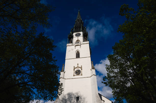 A tall church tower with a clock stands against a bright blue sky, framed by green trees. The Gothic architectural details and the clear weather create a striking and serene image.