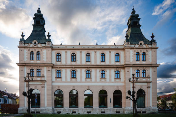 An ornate historic building with detailed architecture and two prominent towers stands against a backdrop of a dramatic sunset sky. The building's symmetrical facade and classic design highlight its grandeur and historical significance.