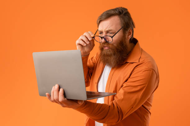 istock Displeased redhaired bearded man looking at laptop through eyeglasses, studio 2164475959