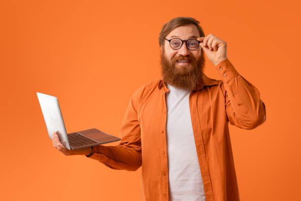 istock Cheerful gingerhead bearded man works on laptop on orange backdrop 2164476017