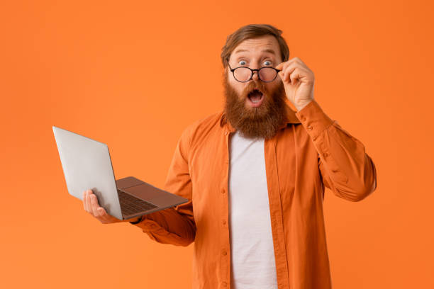 istock Amazed redhaired bearded guy holds laptop looking at camera, studio 2164476024