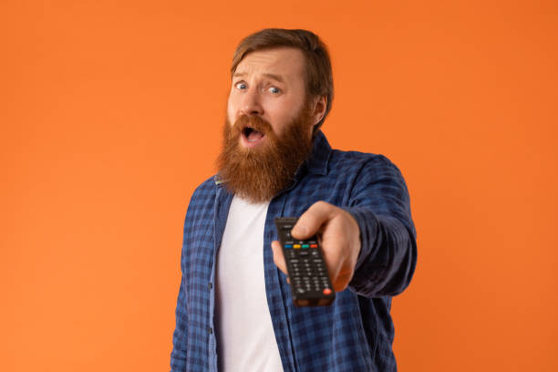 istock emotional bearded guy holding remote control watching tv, orange backdrop 2164476051