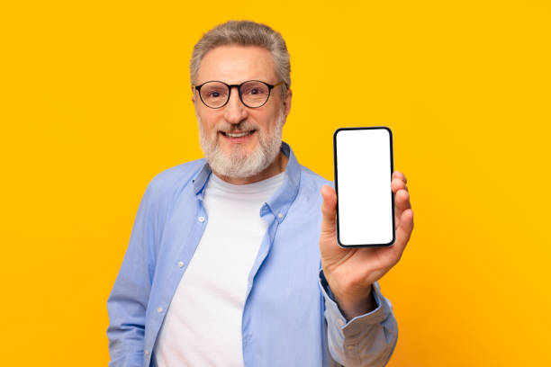 istock smiling senior man showing his smartphone empty screen, yellow backdrop 2164478110