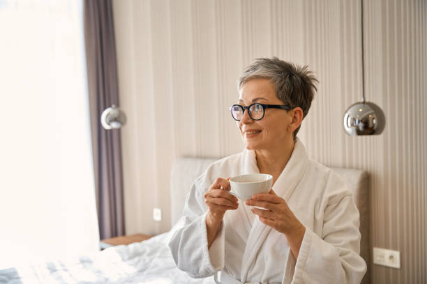 istock Middle age grey-haired woman in bathrobe drinking tea posing in hotel room 2164486843