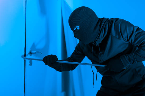 A burglar is shown trying to forcefully enter an apartment at night using a crowbar, illustrating the act of stealing and threatening home safety