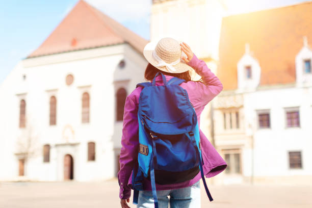 Woman tourist in a straw hat looks at a map in front of historical buildings, signifying exploration and travel, back view