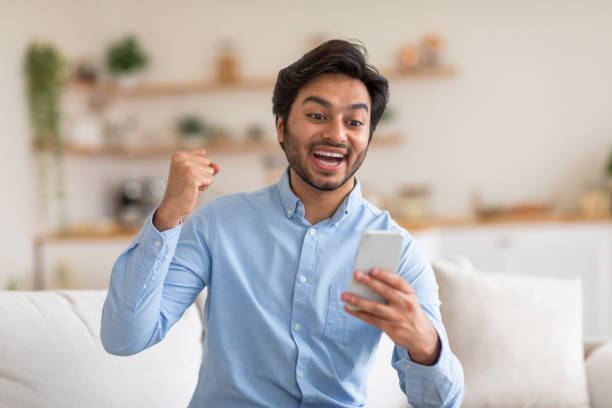 istock Excited Indian Man Sitting on Couch With Phone 2164497660