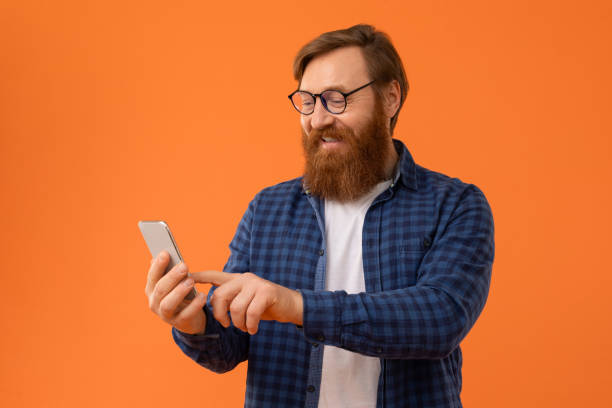 istock redhaired Caucasian man with beard uses his smartphone, orange backdrop 2164497878