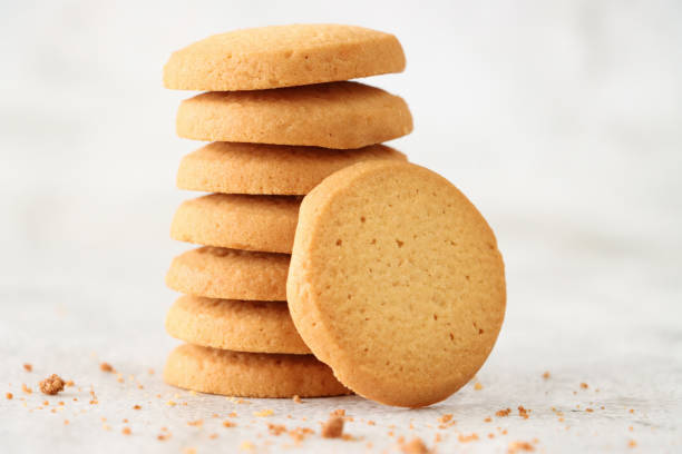 Close-up image of shortbread round leaning against stack of freshly baked, homemade shortcake biscuits surrounded by crumbs, mottled white background, focus on foreground stock photo