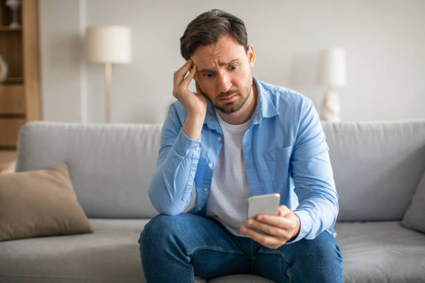 istock Man Sitting on Couch Holding Cell Phone, Touching Head 2164502171