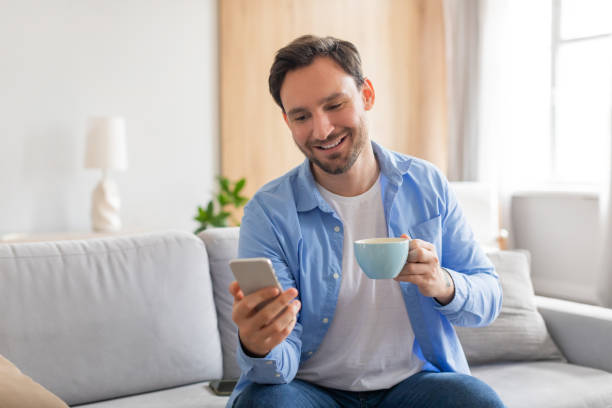 istock Man Sitting on Couch With Coffee and Smart Phone 2164502337