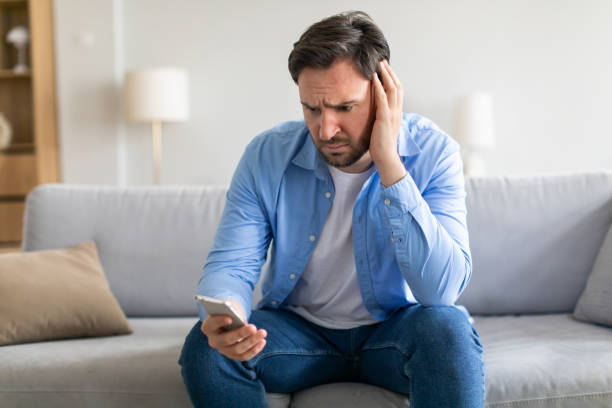istock Frustrated Man Sitting Sofa Holding Head While Looking at Smartphone 2164502350