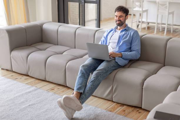 istock Man Using Laptop on Grey Sofa in Modern Living Room 2164505612