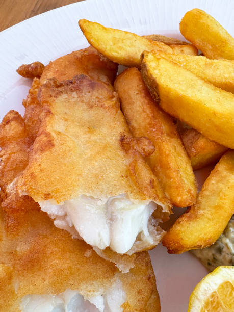 Stock photo  features a plate of crispy, battered cod with golden chips, showcasing tender white flesh fish. A fresh lemon wedge adds a zesty touch, enhancing the dish's flavor. The combination of the crunchy batter, perfectly cooked chips, and vibrant lemon creates a visually appealing and classic presentation