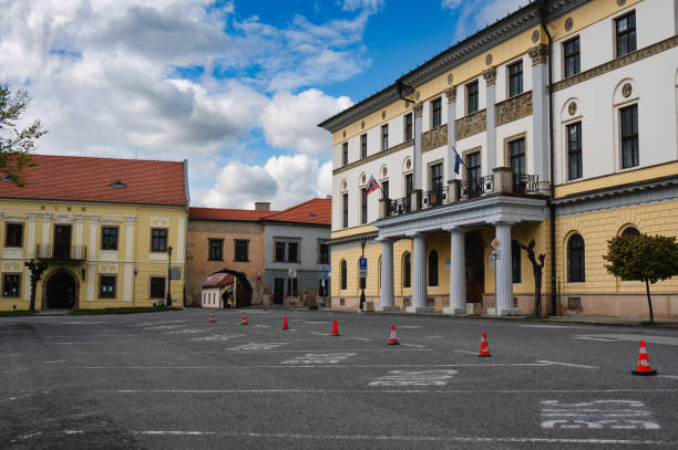 A charming European town street with historic buildings and a cobblestone road under a bright blue sky with scattered clouds. The street is lined with trees and features classic architecture, creating a picturesque and inviting atmosphere.