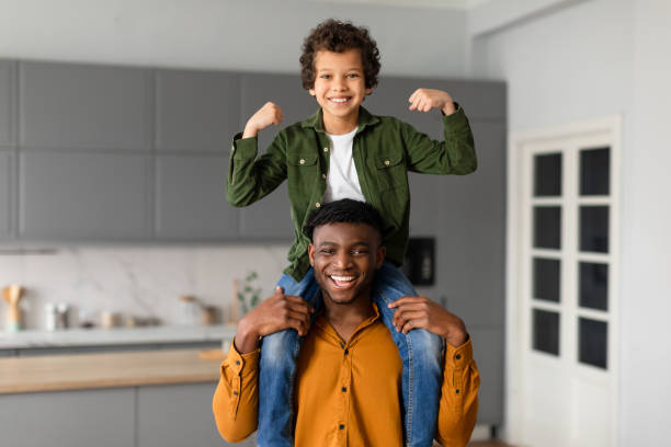 Cheerful African american father carries his child son on his shoulders, boy showing his strength, both smiling in brightly lit, modern kitchen setting at home
