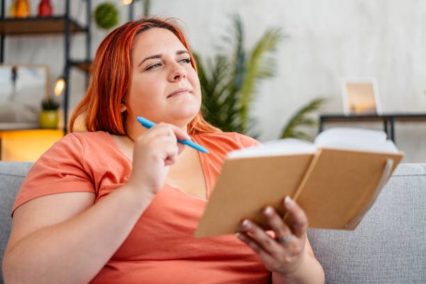 Beautiful young plus size woman writing in a notebook at home.
