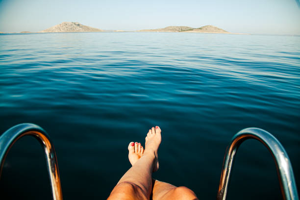 The surreal panoramic island view of Kornati Island National Park in Dalmatia (Croatia) on a clear blue summer day with a bit of morning haze and very calm tranquil sea, the front of a boat in the foreground. From a boat's perspective (boat trip), human feet with legs crossed.