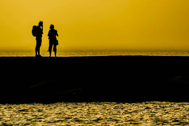 Two people are walking on a pier by the water. The sky is yellow and the water is calm