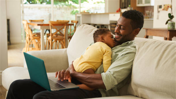 istock Father Comforts Sleeping Baby While Working on Laptop in Bright Living Room 2164606116
