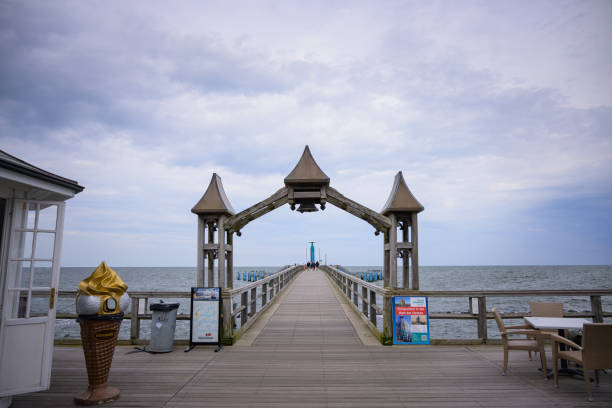 A serene sunset over the sea with a wooden pier reaching into the water. Warm light, soft clouds, and gentle waves create a peaceful, contemplative mood.