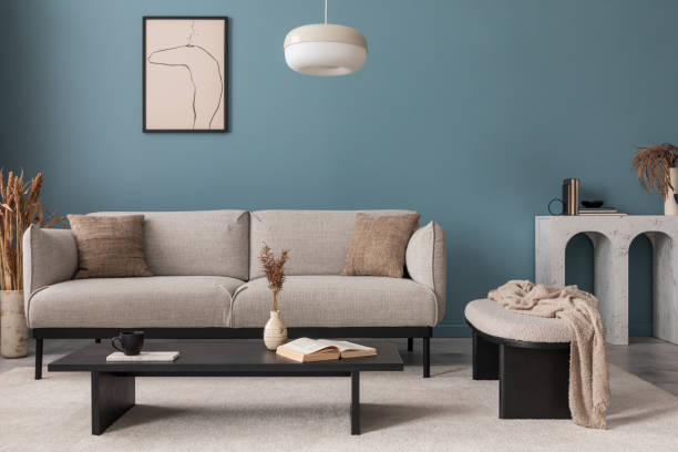istock Warm and cozy blue room with mock up poster frame, cozy sofa, beige bedding, blue wall, pillows, wooden sideboard, bench, gray armchair, vase with flowers plants and personal accessories. Home decor. Template. 2164619151