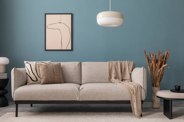 Warm and cozy blue room with mock up poster frame, cozy sofa, beige bedding, blue wall, pillows, wooden sideboard, bench, gray armchair, vase with flowers plants and personal accessories. Home decor. Template.