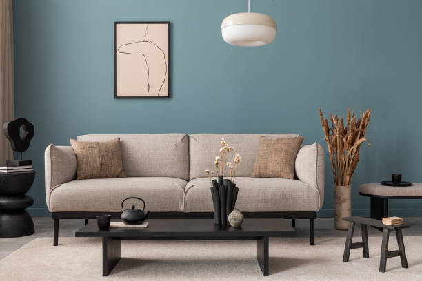 istock Warm and cozy blue room with mock up poster frame, cozy sofa, beige bedding, blue wall, pillows, wooden sideboard, bench, gray armchair, vase with flowers plants and personal accessories. Home decor. Template. 2164619157