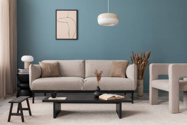 istock Warm and cozy blue room with mock up poster frame, cozy sofa, beige bedding, blue wall, pillows, wooden sideboard, bench, gray armchair, vase with flowers plants and personal accessories. Home decor. Template. 2164619205