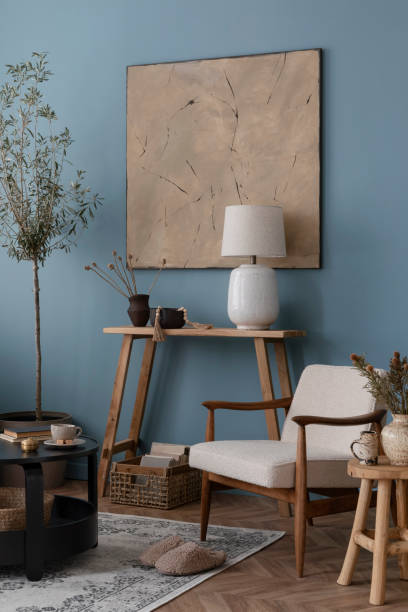 istock Warm and cozy blue room with mock up poster frame, cozy sofa, beige bedding, blue wall, pillows, wooden sideboard, bench, gray armchair, vase with flowers plants and personal accessories. Home decor. Template. 2164626514