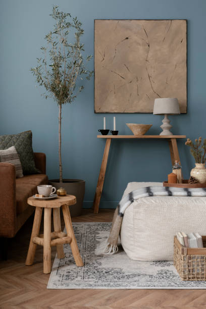 istock Warm and cozy blue room with mock up poster frame, cozy sofa, beige bedding, blue wall, pillows, wooden sideboard, bench, gray armchair, vase with flowers plants and personal accessories. Home decor. Template. 2164626537