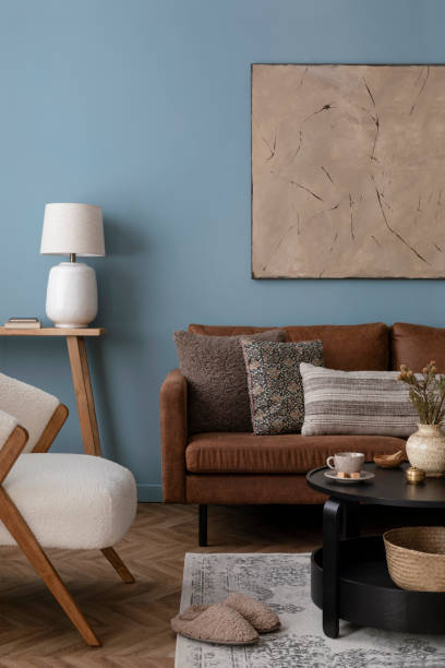 istock Warm and cozy blue room with mock up poster frame, cozy sofa, beige bedding, blue wall, pillows, wooden sideboard, bench, gray armchair, vase with flowers plants and personal accessories. Home decor. Template. 2164626559