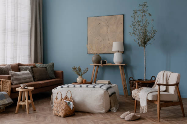 istock Warm and cozy blue room with mock up poster frame, cozy sofa, beige bedding, blue wall, pillows, wooden sideboard, bench, gray armchair, vase with flowers plants and personal accessories. Home decor. Template. 2164626565