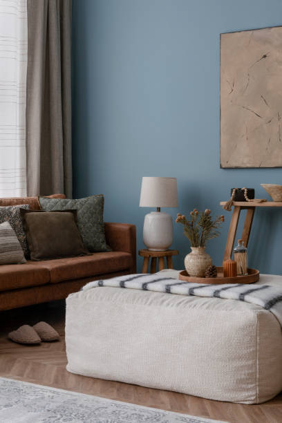istock Warm and cozy blue room with mock up poster frame, cozy sofa, beige bedding, blue wall, pillows, wooden sideboard, bench, gray armchair, vase with flowers plants and personal accessories. Home decor. Template. 2164626568