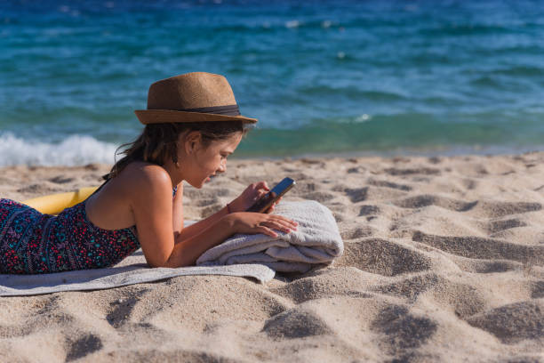 Teenage girl wearing a sun hat and lying down on a beach and using smart phone
