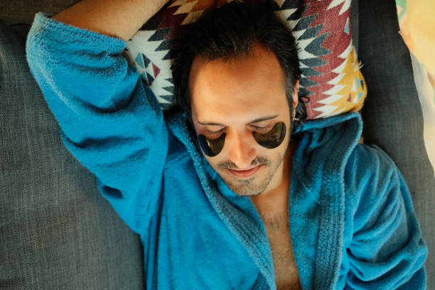 istock Man relaxing on couch wearing a bathrobe and using cosmetic eye patches 2164636932