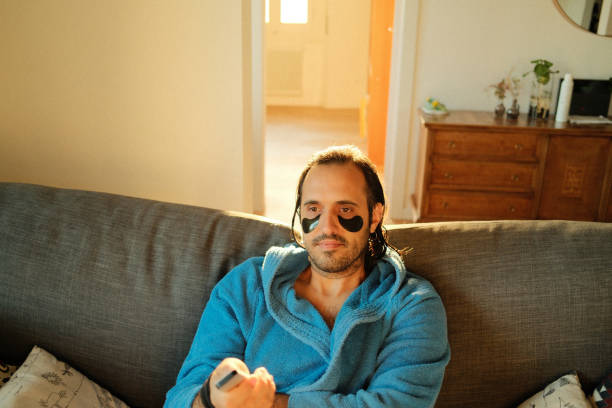 Front view of handsome peaceful African American bearded man with white facial mask, sitting in lotus position on sofa with laptop, meditating with happy smile