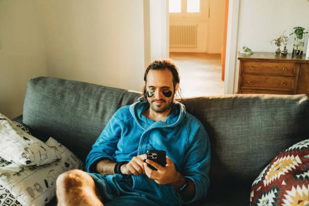 istock Man relaxing on sofa at home using smartphone with eye patches 2164636968
