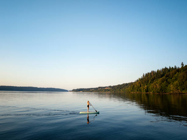 A woman enjoys paddleboarding in Washington state, USA on a beautiful summer morning. The rising sun glows along the horizon. One of the many ways to enjoy the beauty of the PNW.  Shot on the Puget Sound.