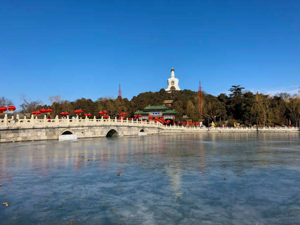 A serene view of the White Dagoba standing tall on Jade Flower Island in Beihai Park, Beijing, during the Spring Festival. The frozen lake in the foreground reflects the clear blue sky and traditional Chinese architecture adorned with festive red lanterns, capturing the essence of winter tranquility and cultural celebration.