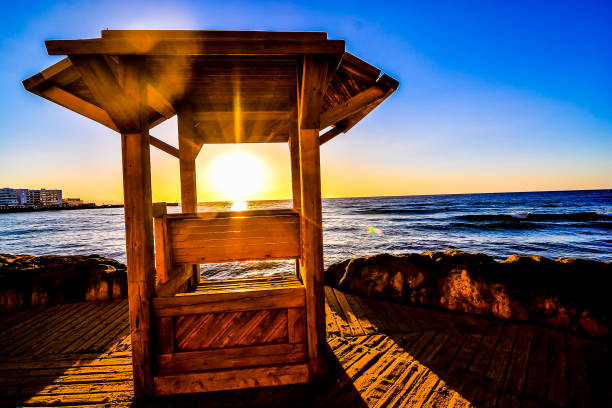 A wooden structure with a roof is sitting on a beach. The sun is setting and the sky is blue