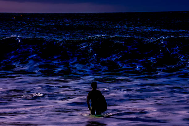 A surfer is standing on a surfboard in the ocean at night. The water is dark blue and the sky is cloudy