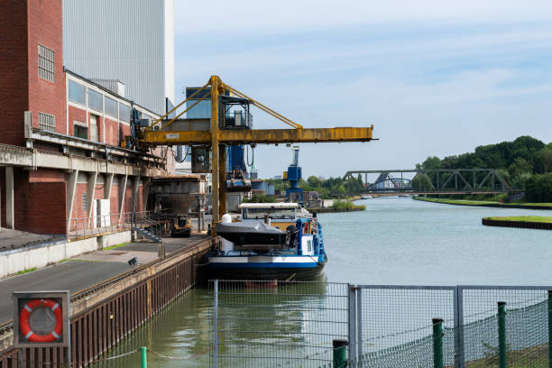 A crane operates at an industrial dock, transferring cargo onto a moored boat along the river on a clear, sunny day.