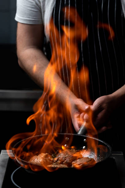 Cooking by a chef, processing food with a flame in a frying pan on a black background, the concept of restaurant service, impressive cooking