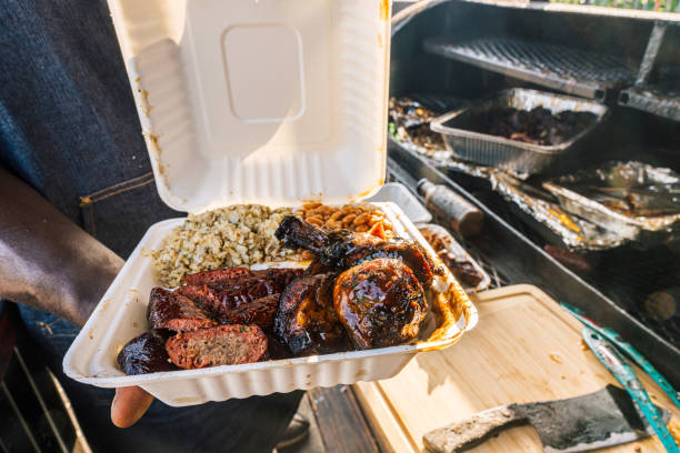 istock Close-Up BBQ Chef holding open a Compostabale Takeout Container full of Smoked BBQ Beans, Sausage, Chicken Thighs & Drumsticks, and Dirty Rice from a Food Truck or Traveling Smoker Catering Company 2164703087