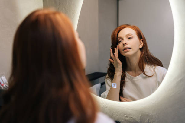 istock Reflection on mirror of attractive young redhead woman enjoying her appearance while getting ready in bathroom. Happy beautiful female touching face look 2164712326