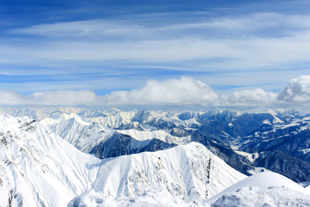 View of snow-covered mountain peaks over which the clouds are floating. In the background is a blue cloudy sky