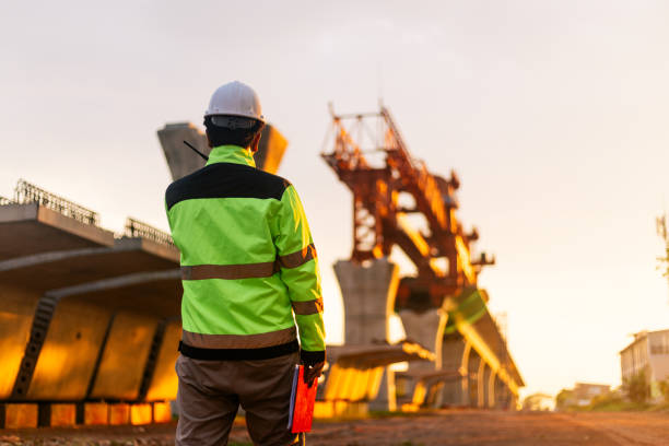 istock A construction worker is standing on a bridge, talking on his cell phone. The scene is set in a construction site, with a large crane in the background. The worker is wearing a yellow jacket 2164722057